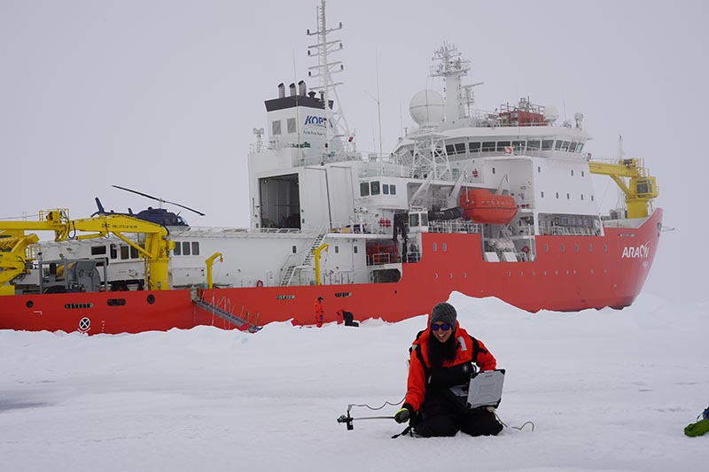 Alia Khan kneeling in the snow with a computer in her lap and holding a scientific instrument on a stick. She is wearing a bright red snow coat, which matches the bright red paint on the large ship behind her.