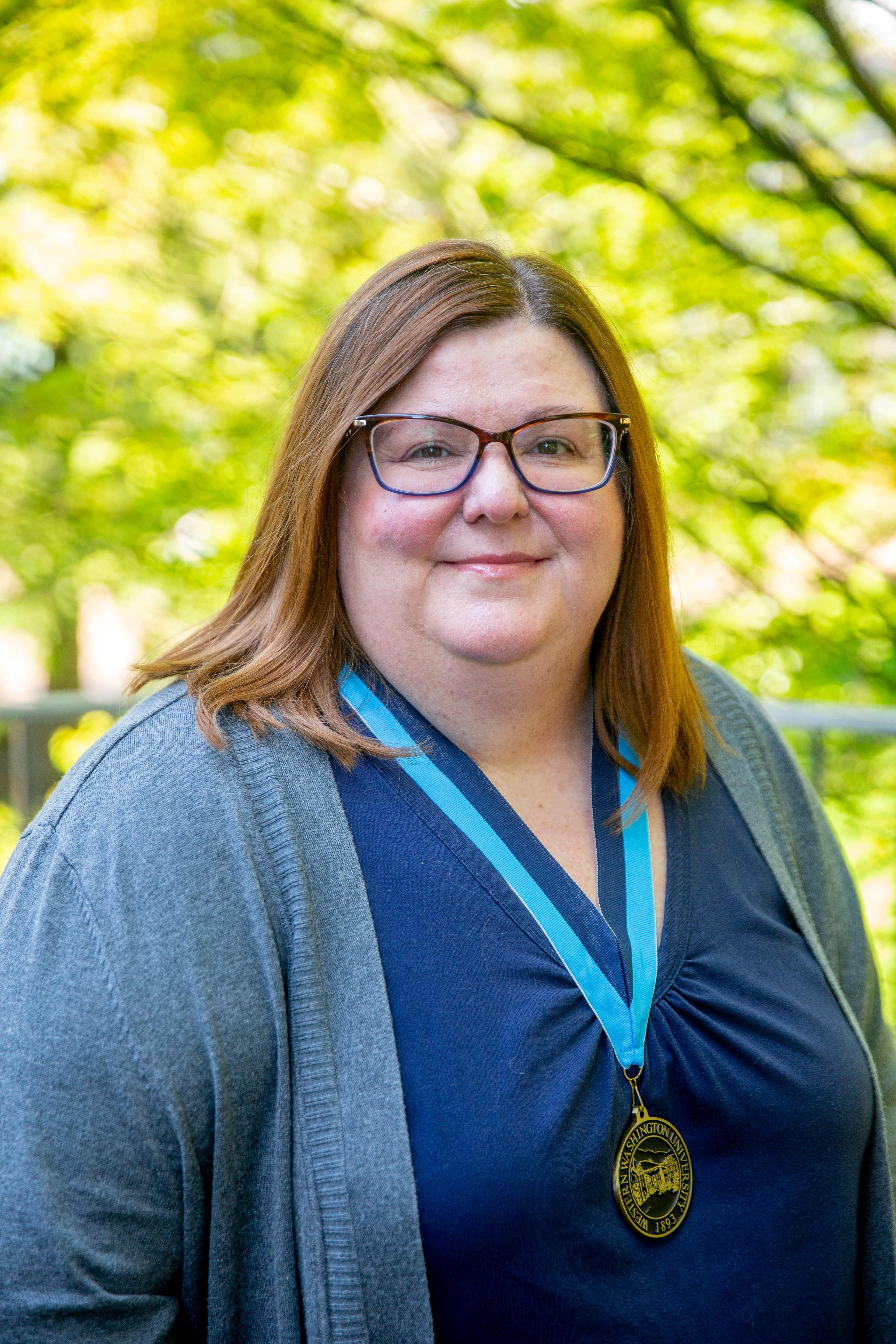Carrie Arrnett smiles wearing a WWU medallion on a neck ribbon with sun dappled leaves in the background