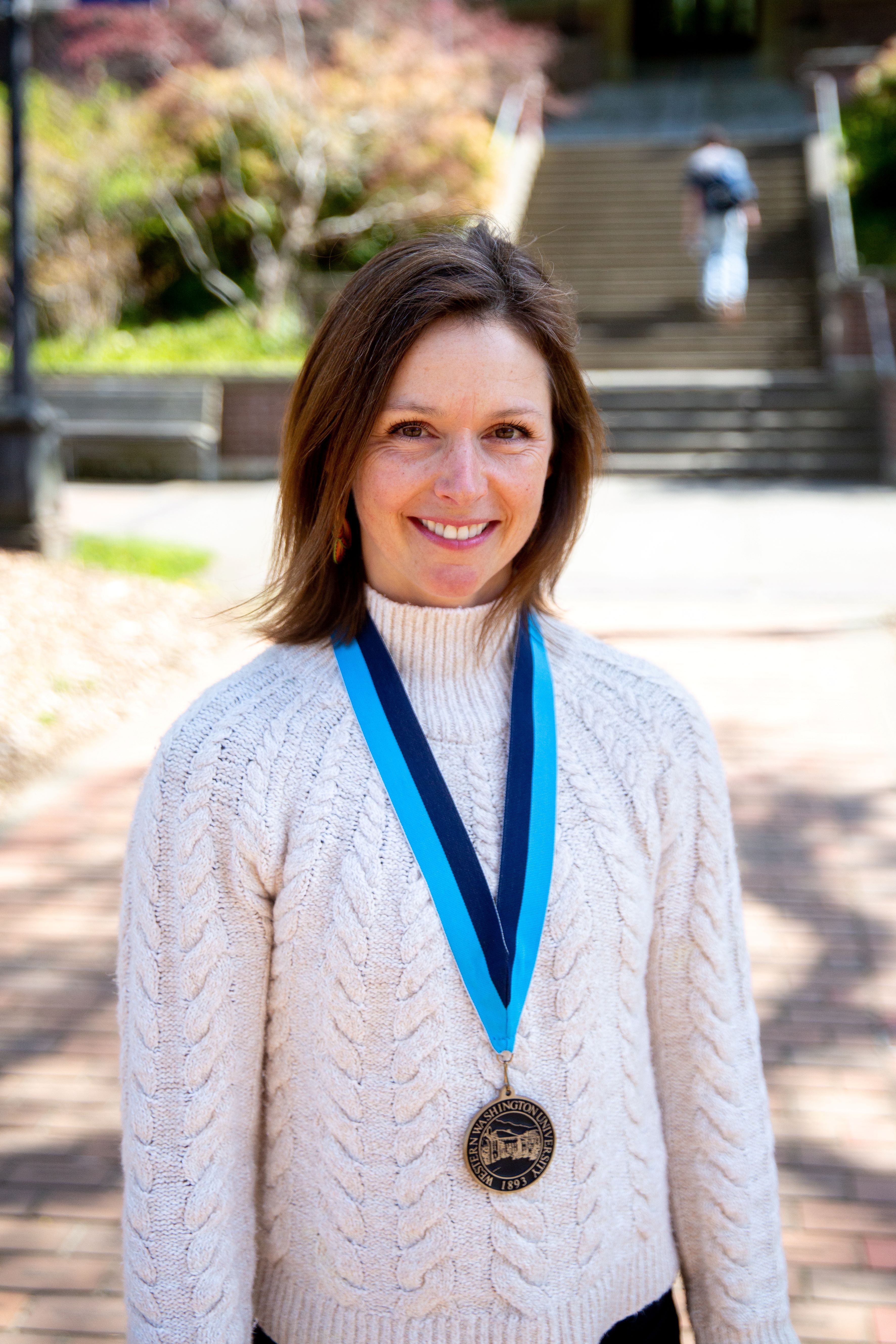 Kate Stevenson in a white cable knit sweater standing in front of Old Main with a WWU award medallion on a neck ribbon