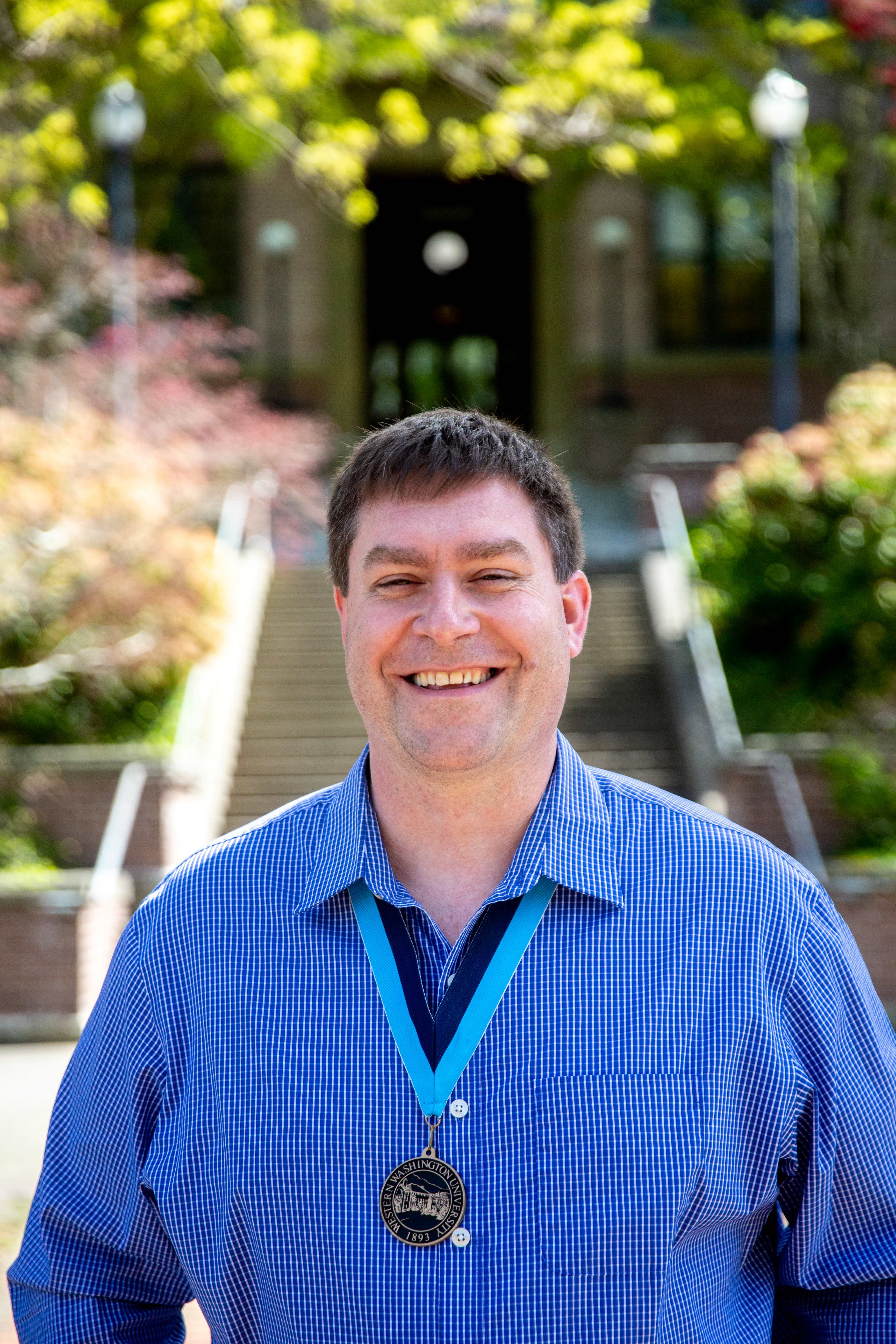 Ryan Cullup with a broad smile in front of Old Main wearing a vivid blue shirt and a WWU medallion on a neck ribbon 