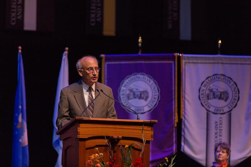 Sabah in a dark beige suit jacket and tan striped tie addresses the convocation attendees. The college flags and banners are behind Sabah.