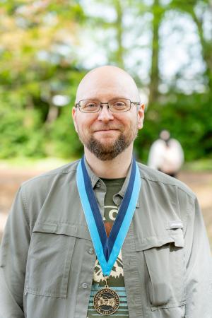 Zach McGrew smiling and wearing a WWU medallion award on a neck ribbon with trees in the background