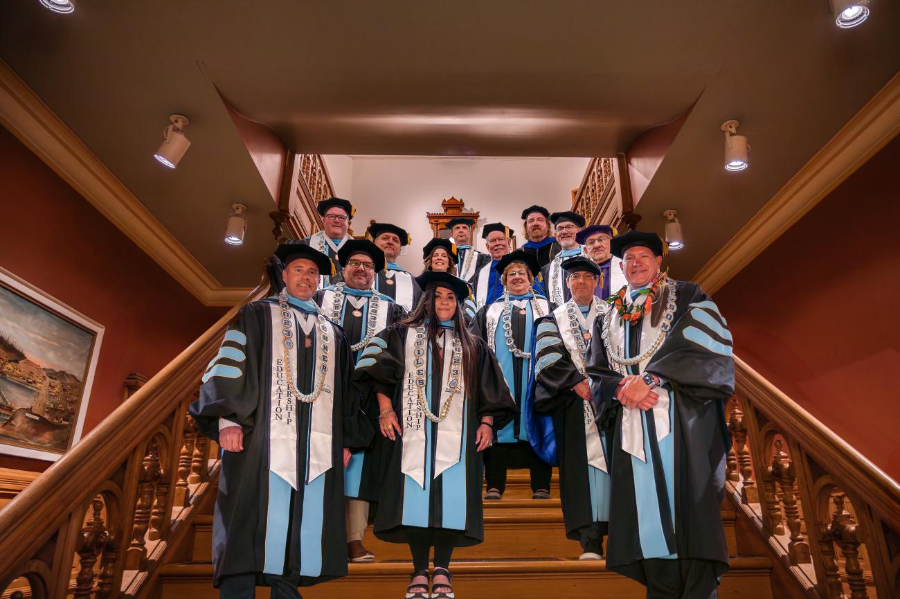 several doctoral graduates and faculty wearing academic regalia smile for the camera on an ornate stairwell
