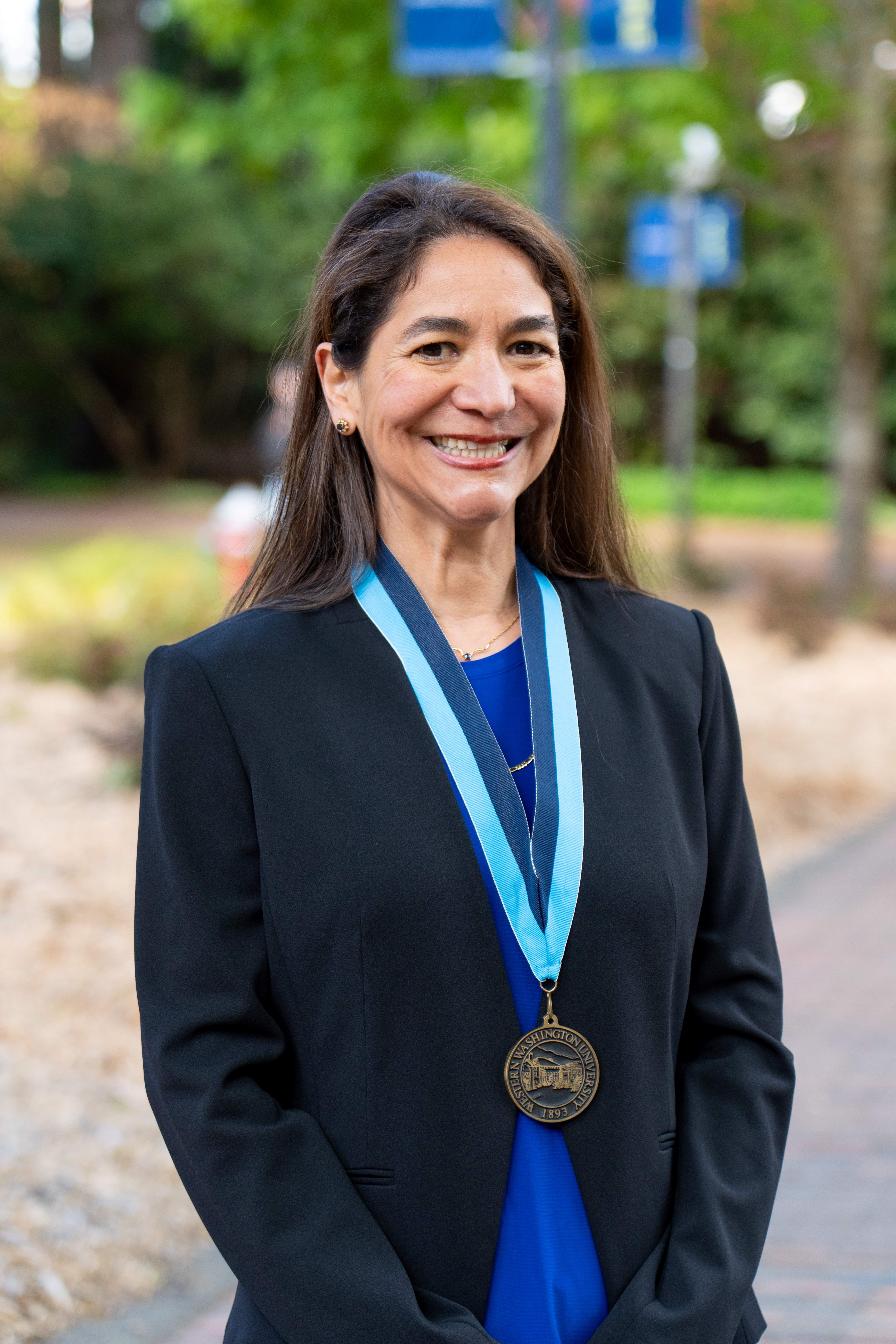 Dr. Liliana Deck stands in front of Old Main with a broad smile and wearing a WWU award medallion