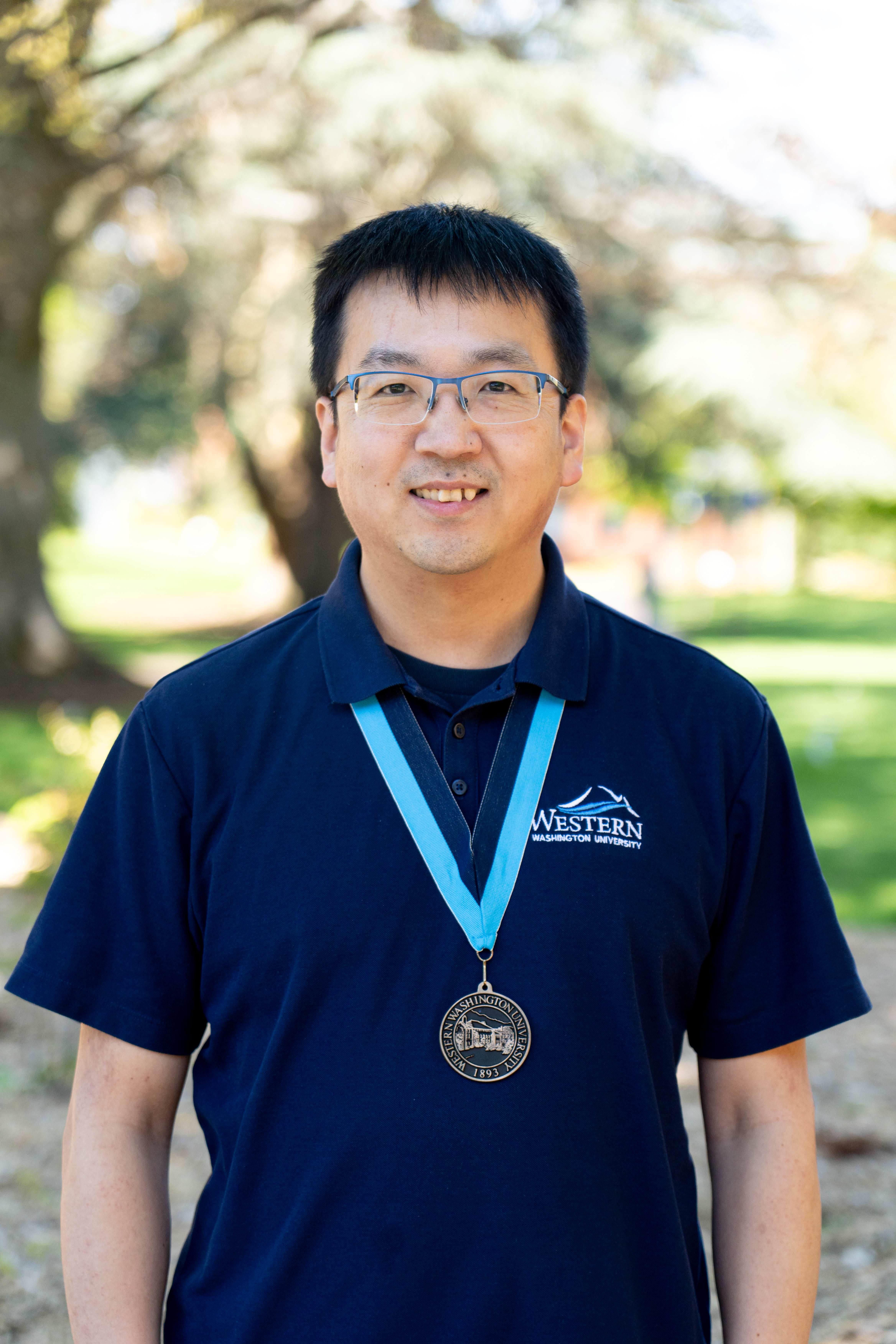 Dr. Shuo Li stands in front of Old Main wearing a WWU polo shirt and an award medallion